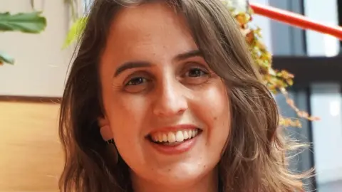 Lucy Jenkins Head shot of Lucy Jenkins, a white woman in her thirties. She has shoulder length layered light brown hair, is wearing light make-up and dangly earrings and is smiling broadly at the camera. She appears to be indoors in a workplace.