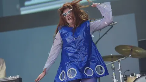 PA Media Musician CMAT in a blue dress dancing on stage in front of a drum set.