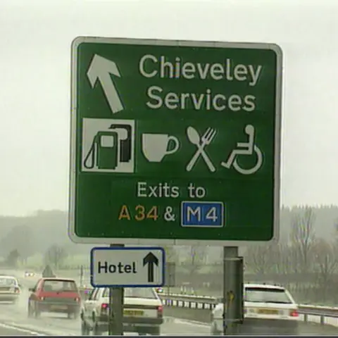 A green motorway Chieveley Services sign with white images and lettering on the carriageway where cars can also be seen. 