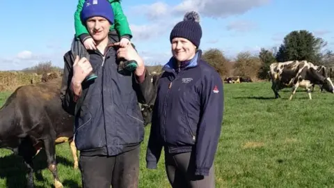 Staffordshire County Council Richard and Ella Upton are stood in a field with three-year-old son Reg on Richard's shoulders. All three are wearing outdoor coats and bobble hats. There are cows behind them.