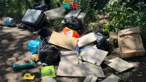 Jennifer Wells Rubbish, including what could be a paint bucket on the far left and a grey suitcase, is near bushes. Cardboard and black bags are in the foreground. 