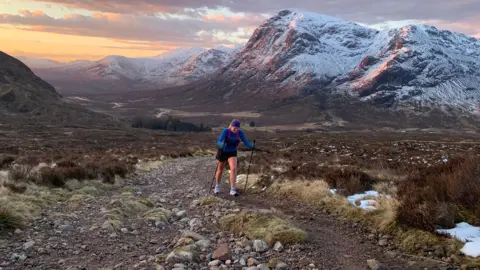 Freya Shepherd Freya running on the mountain trail with poles in her hands. It is dusk and the mountain tops covered in snow behind her are lit with warm orange light.
