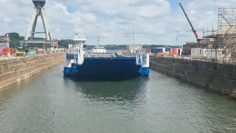 Tamar Crossings The vessel Lynher in the dry dock, except the dry dock is filled with water. The boat is blue and white and is tied to the sides of the dock. There is scaffolding on the dock on the right-hand side and a crane. The water is a murky blue. There are clouds in the sky.