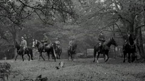 Black and white image of 6 riders on horses wearing riding gear and flat caps.  They are framed in the image by tree branches overhead.