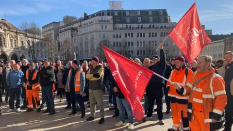 Protestors gathered outside Birmingham council house. Two people are each holding a red "Unite the Union" flag. The sky is blue in the background.