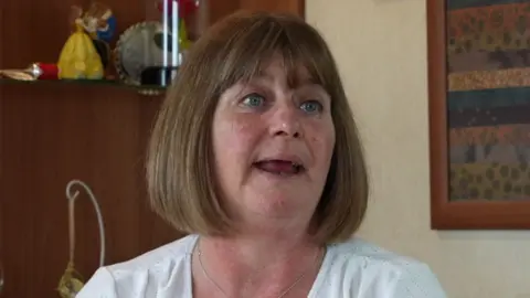 Mandy Dossett, a woman in her 50s, sits in what appears to be the lounge of a home. Her brown hair is in a shoulder-length bob and she is wearing a short-sleeved white blouse.
