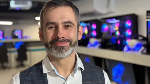 Ian Irving is smiling at the camera. He has grey hair and beard and brown eyes. He is wearing a white shirt and a waistcoat. 