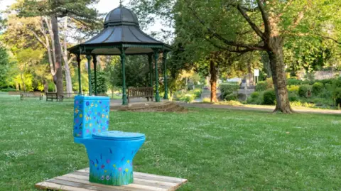 Sienna Anderson A blue toilet patterned with colourful flowers. A bandstand is in the background.