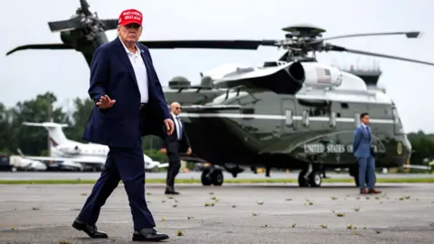 Trump, wearing a red MAGA cap, walks infront of the Marine Force One helicopter, and waves.
