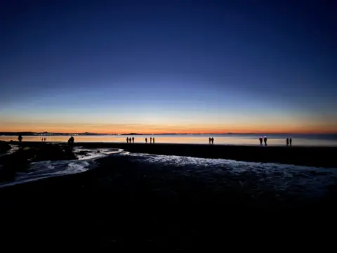 Eleanor Ammons A dark beach. There are people standing at the water's edge and the horizon is orange.