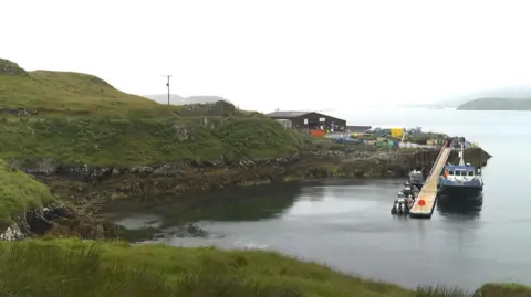 A brown building is on a small promontory of land sticking out into the sea. The sea is flat calm, and it is a hazy day. There is a small jetty with a small boat tied up to it. The boat is white and blue in colour.