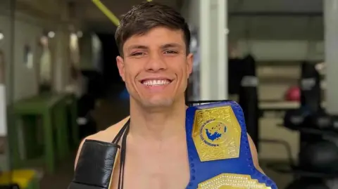 A man with dark hair smiling with a boxing belt over his shoulder.