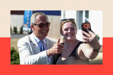 Reuters Reform UK party leader Nigel Farage poses for a selfie with a woman on the day of the Clacton by-election. He is wearing sunglasses, a blue and white striped blazer, a white shirt and blue tie, and is giving a thumbs up gesture. The woman holds the phone with her outstretched arm.