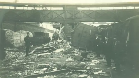 Fred Spalding A black and white photograph showing the aftermath of the Witham railway crash. A carriage is turned on its side and is lying on the platform, next to upright carriages. The platform is covered in debris. There is a bridge over the railway tracks, and people are stood on the bridge as well as on the platform.