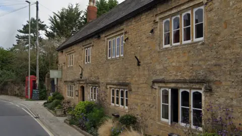 Google Google Street View of the Ollerod. A red phone box stands on the pavement near the entrance which is surrounded by green shrubs, ornamental grasses and purple-flowering plants.