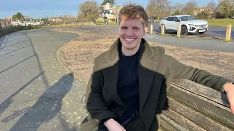 A photo of James Vincent sitting on a bench in the sun, wearing a dark green coat. In the background is trees, a road and a parked car. 