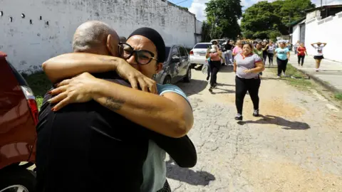 Reuters Two people are seen embracing tightly on a street in Tocuyito, Venezuela, with a group of people walking in the background and cars parked along the road.