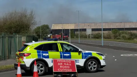 A police car in front of the same building engulfed in smoke,. the care has a sign in from on the ground in red and white saying road closed the welsh equivalent