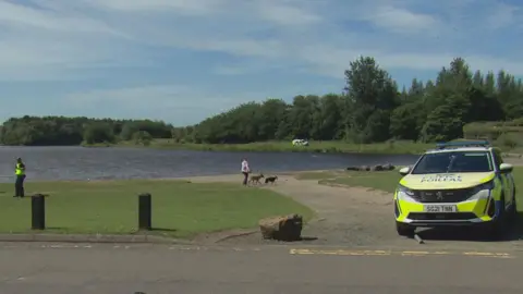a police are is parked next to the loch. A police officer and a woman walking two dogs are on a path