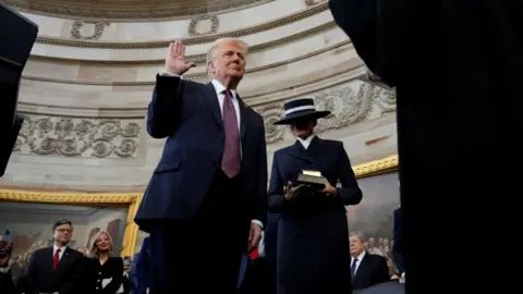 Reuters Trump angle from the bottom down with hand raised as he takes oath of office. Melania Trump just behind him holding Bible