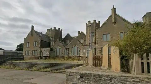 There is an old Victorian building with some interesting architectural features. It looks neglected. Weeds are growing on the steps leading to the building and spreading across the paving stones. There is a small monument in the foreground at the right hand side. 