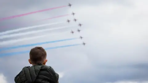 A young boy, seen from behind, looking up at the sky as nine aeroplanes fly through it. Behind them they are leaving red, white and blue vapour trails.