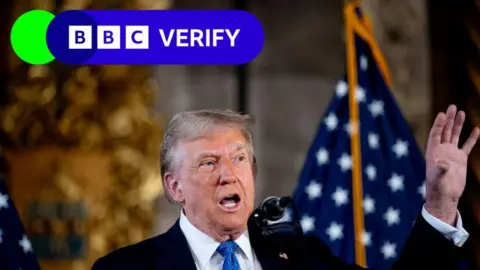 Donald Trump speaking at a conference. He is wearing a suit and tie, with his left arm raised.  