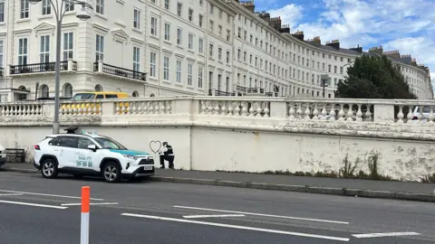 George Carden/BBC The mural on a wall with the regency buildings of Palmeira Square behind. A taxi is parked next to it