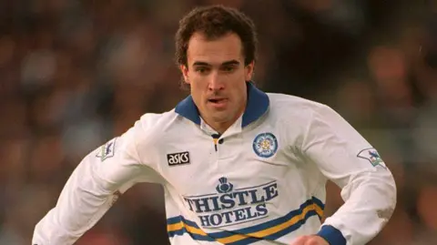 Getty Images A male footballer with short brown hair runs with his tongue sticking out his mouth. He is wearing a white, blue and yellow Leeds United shirt.