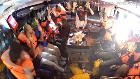 Activists in orange life jackets holding up their hands onboard a boat being boarded by Israeli forces