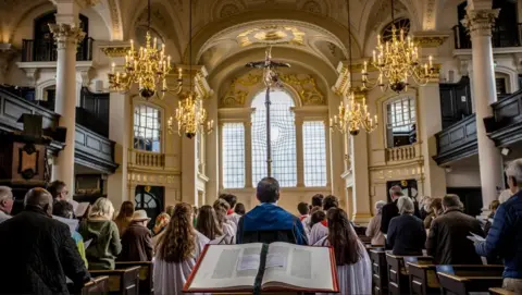A service taking place inside St Martin-in-the-Fields with people in the pews standing and a book in the middle of the church with a choir of people facing away from the camera