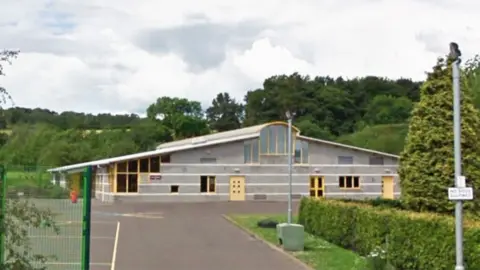 Google A grey primary school with yellow windows and doors, surrounded by trees