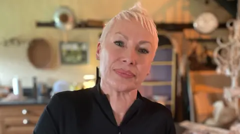 A head and shoulders image of a woman with very short peroxide blonde hair and wearing a black shirt, with a blurred background of a kitchen