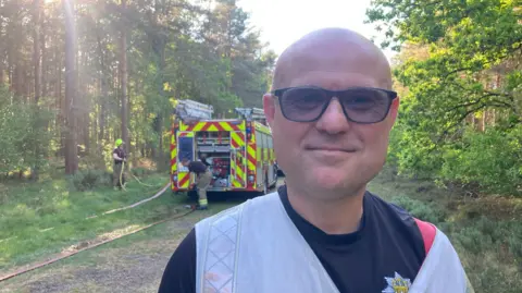 Mr Reeve is standing in the forest in front of a fire engine. He is smiling at the camera while wearing sunglasses. He is wearing a blue top - with the fire service logo on the breast. Behind him two firefighters are taking equipment out of the engine. 