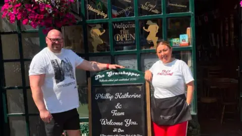 Katherine Harvey Katherine and Phillip Harvey stand outside The Grasshopper pub, smiling to the camera. 