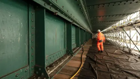 Gateshead Council Close-up view of an old section of the Tyne Bridge showing scaffolding and the structure's heavily riveted lower west arch, which is rusty and has a colour in the shade of Hollybush Green. There is a worker in orange uniform walking away on the scaffolding.
