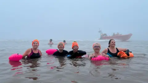 Five women are in the sea smiling at the camera. Four of them are wearing fluorescent hats and all of them have floats. It is foggy. There are two men swimming and a rigid inflatable boat driving behind them.