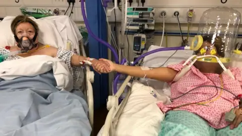 A mother and daughter in intensive care in beds next to each other. Maria Rico, is wearing a mask over her face to help her breathe while in a hospital bed while her daughter Anabel Sharma is in the bed on the right wearing a helmet-like covering over her head attached to an oxygen pump.