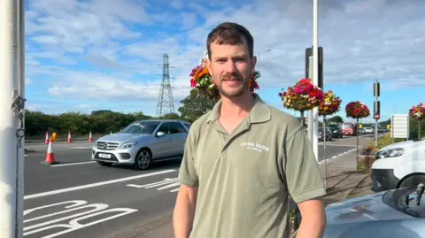 A man is wearing a green polo looling at the camera, and behind him is a car driving on a carriageway.
