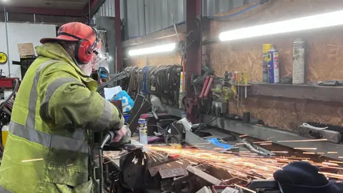 Person wearing protective gear uses an angle grinder on metal in a workshop, with bright sparks flying across a workbench cluttered with tools and equipment