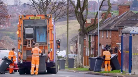 Three refuse collectors wearing orange hi-vis uniform bringing bins from a street to a refuse truck