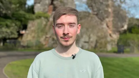 Rob Trigg/BBC Rees is photographed looking into the camera. He is wearing a sea-green jumper, and is stood in front of the Bridgnorth castle ruins. The sky is blue and there's strong sunshine. 
