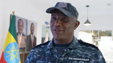 A head and shoulders image of a man in a blue camouflage uniform and peaked cap. A badge says "Eth Federal Police" on his chest and an Ethiopian flag is in the background.