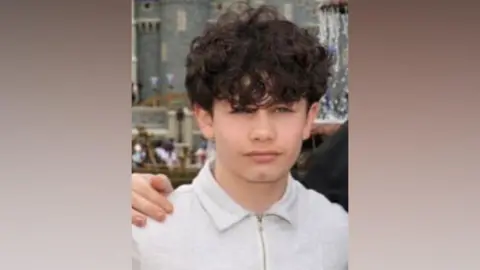 A young boy wearing a white polo shirt. He has curly brown hair. Someone has their hand on his shoulder. Behind, out of focus in the background, is a medieval castle-looking building.