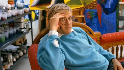 David Hockney in his Studio in California in 1987 wearing a blue jumper relaxing on a chair with his hand to his face 