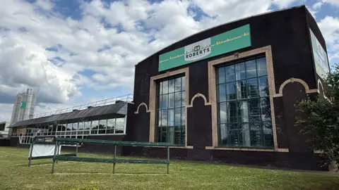 BBC/Chris Long The Roberts Bakery buildings, showing a dark-brown brick-built building with beige bricks bordering the windows and a lower window-fronted shed with three silver silos behind it