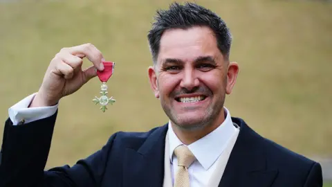 PA Media Head and shoulders of Jordan Wylie after being made an MBE at Windsor Castle. He is standing in the gardens, which are blurred, and is wearing a dark suit, white shirt and lemon tie holding his cross-shaped medal with a red ribbon in his right hand next to his face. He has dork spiky hair with flecks of grey, stubble, and is grinning