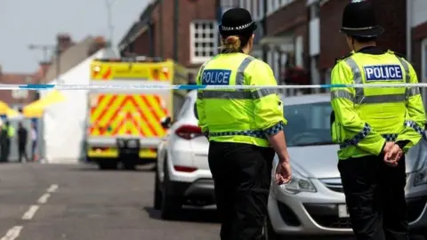 Two police officers stood behind some police tape. In the background is a white police tent and other emergency service workers. 