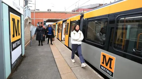 Passengers walk along a Metro platform at Millfield station next to the new train. It has a yellow-and-grey livery with the Metro logo on the side of one of the carriages.