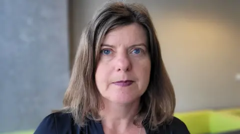 Sandy Brindley, who has blue eyes, sandy-coloured hair in a long bob and purple lipstick, looks into the camera